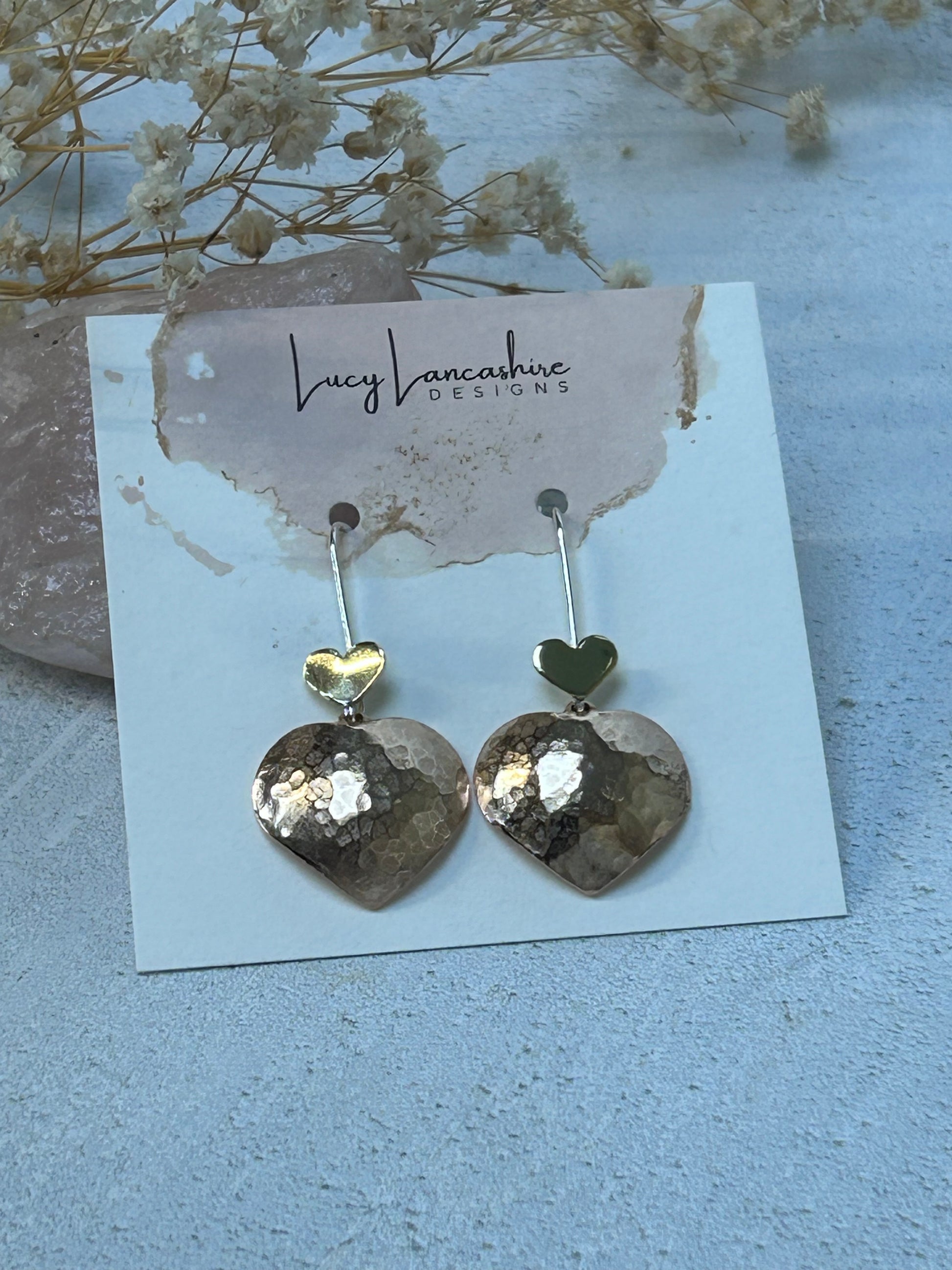 Pair of silver earrings on a card with 'Lucy Lancashire Designs' branding, placed on a light surface with dried flowers.